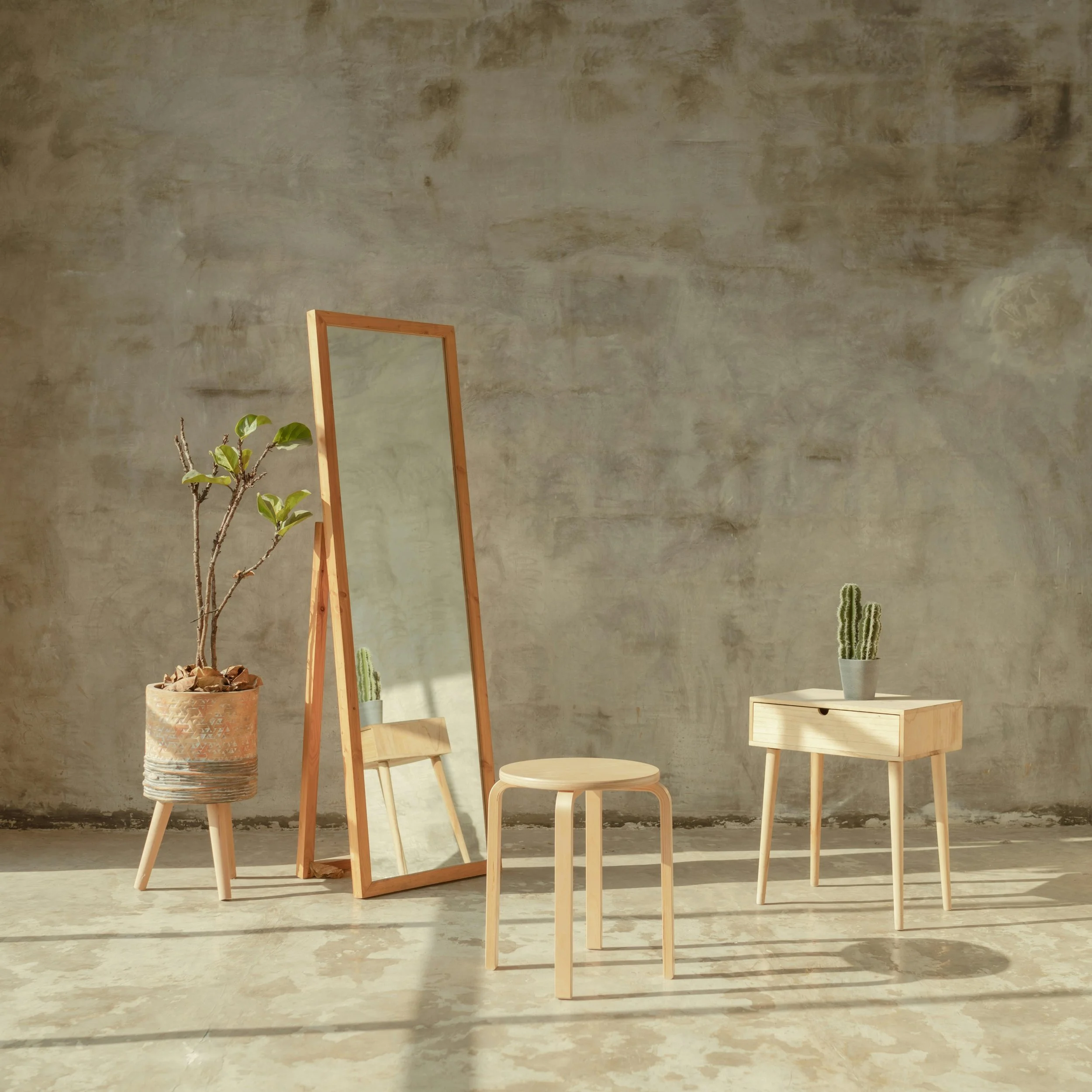 Minimalist interior with a large textured concrete wall, wooden furniture including a small table, a stool, a mirror frame, and potted plants on the floor and on a small wooden drawer.