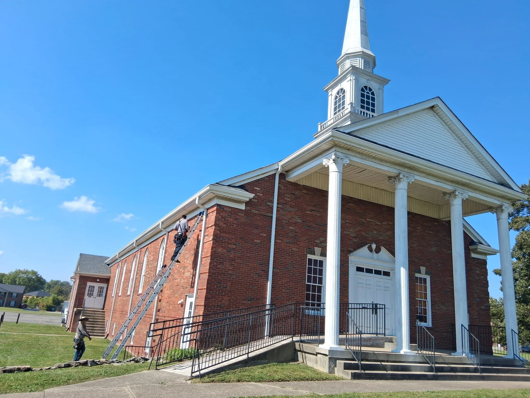 A man working on the exterior of a brick church building with white columns and a steeple, on a sunny day with a blue sky and few clouds.
