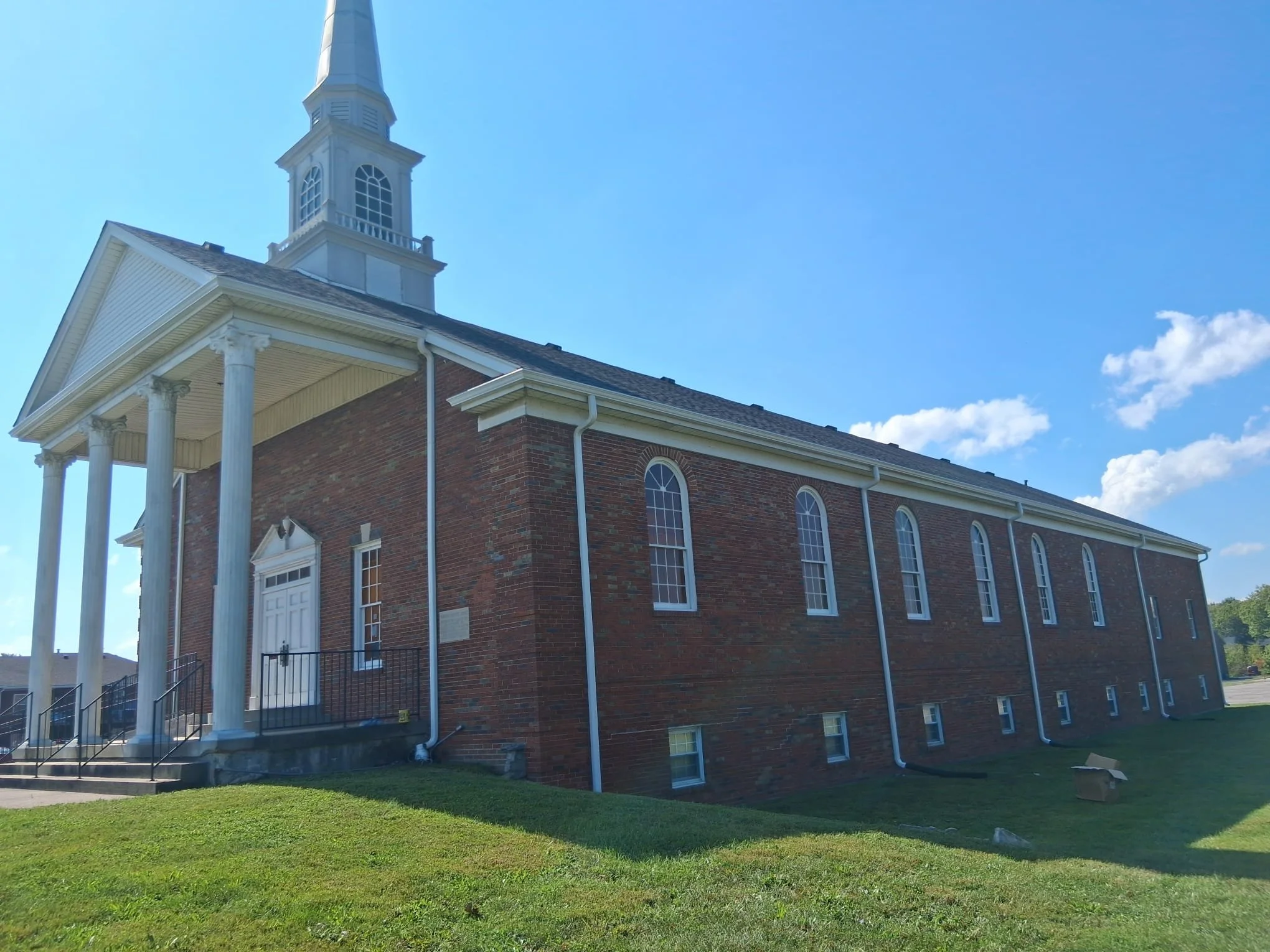 A church building with a brick exterior, tall arched windows, white columns at the entrance, and a steeple on the roof, set against a blue sky with some clouds.