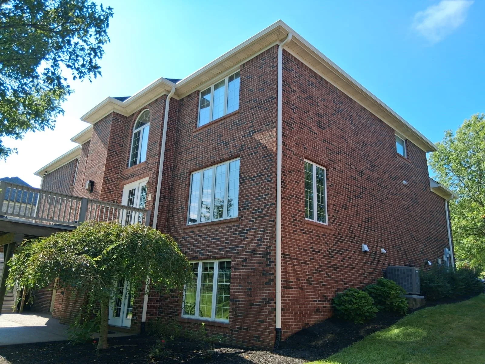 A three-story brick house with multiple windows and a wooden deck. There are trees and green bushes around the house, with clear blue skies overhead.