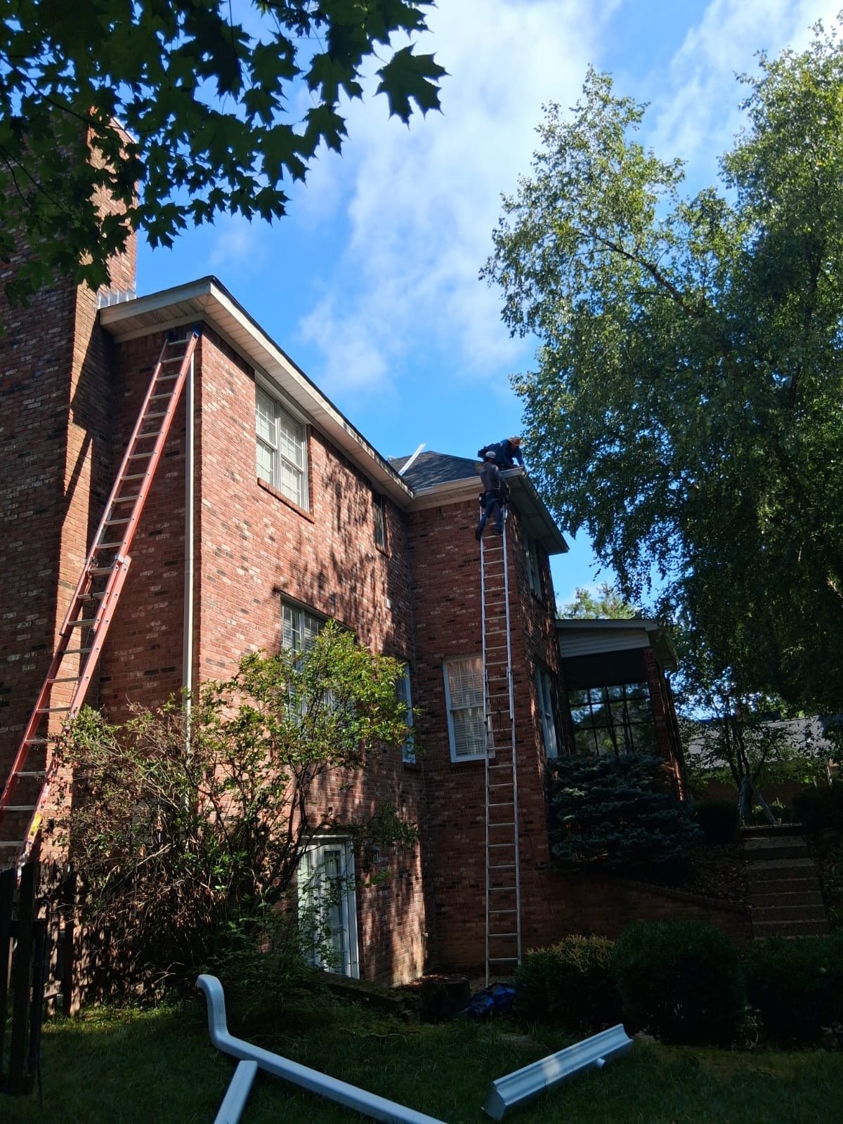 Two workers on ladders repairing the roof of a multi-story brick house during daytime. The house is surrounded by green trees and bushes, and the sky is partly cloudy.