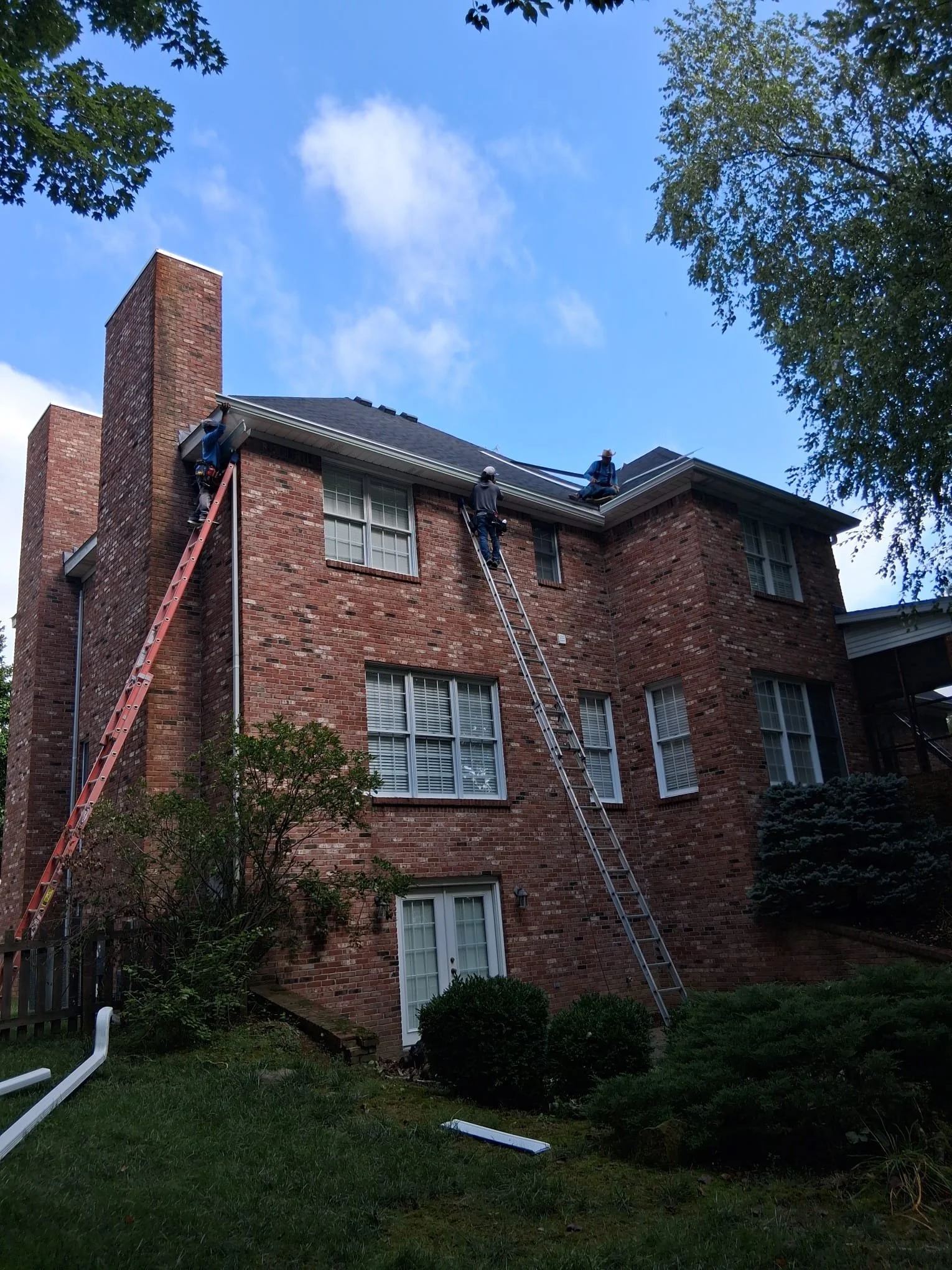 Three workers on ladders repairing or inspecting the roof of a brick house, with a blue sky and some trees in the background.