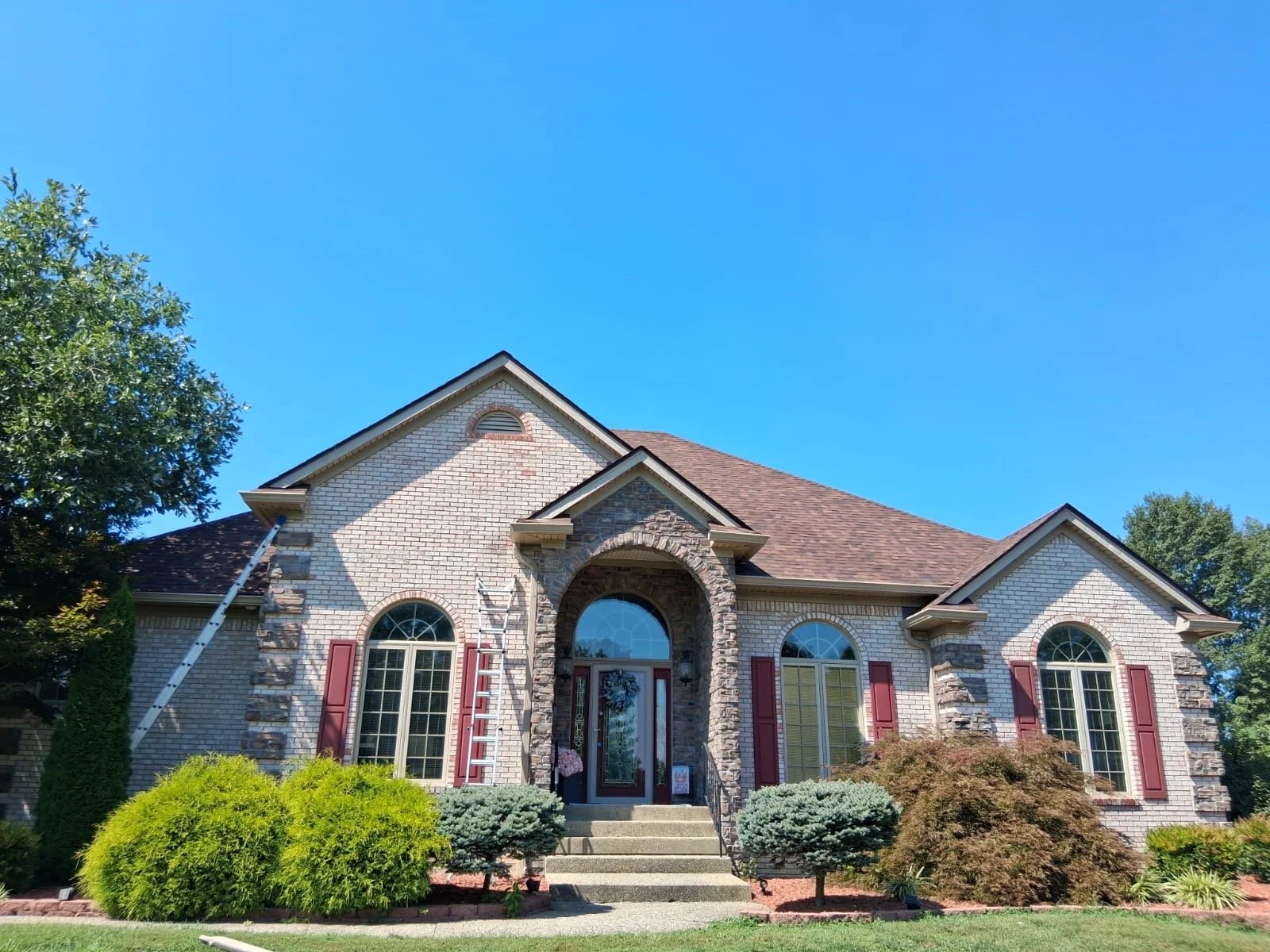 Large brick house with arched windows and red shutters, surrounded by landscaped bushes and trees, under a clear blue sky.