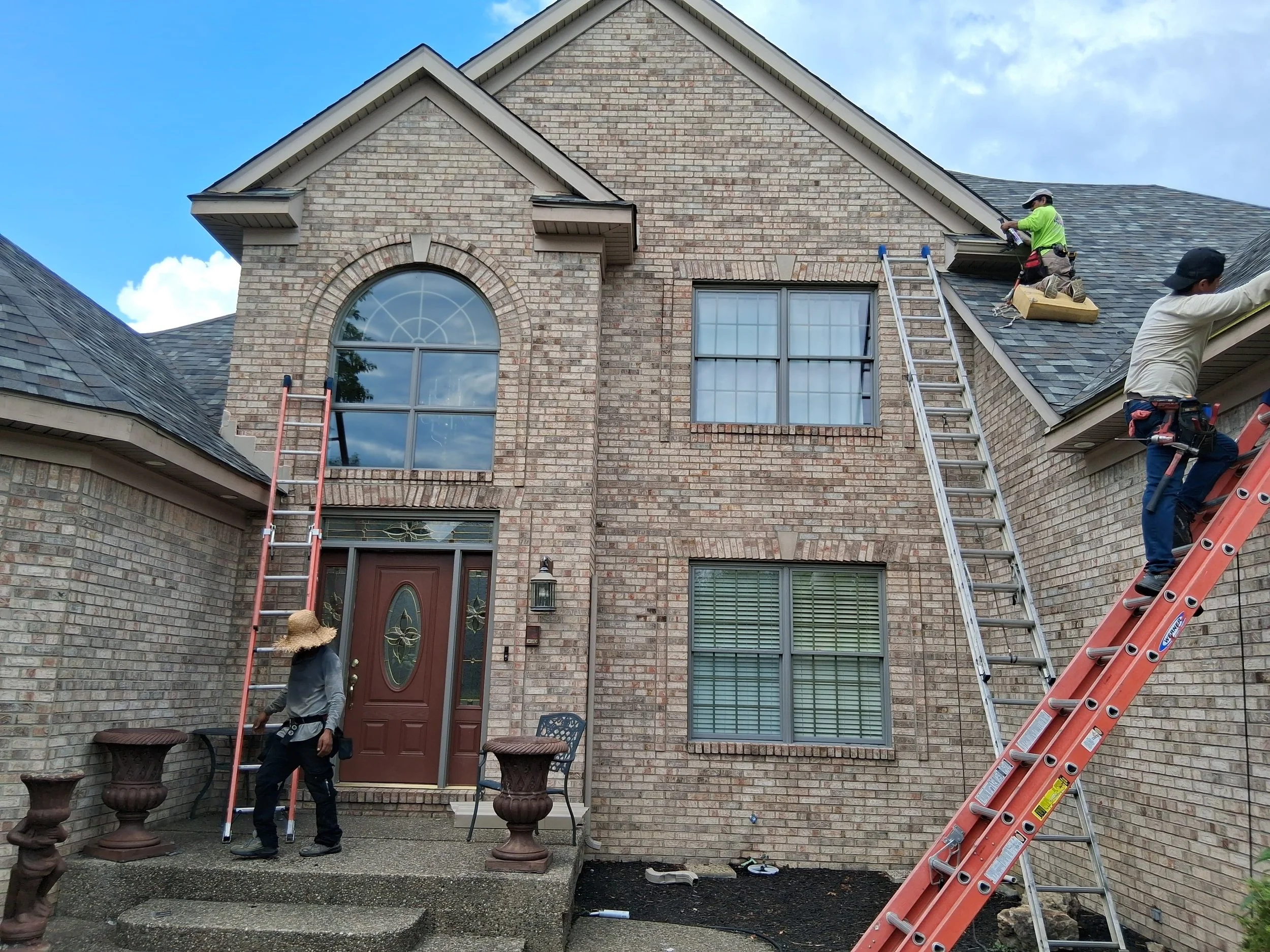 Workers installing or repairing roofing on a brick house with ladders and tools.