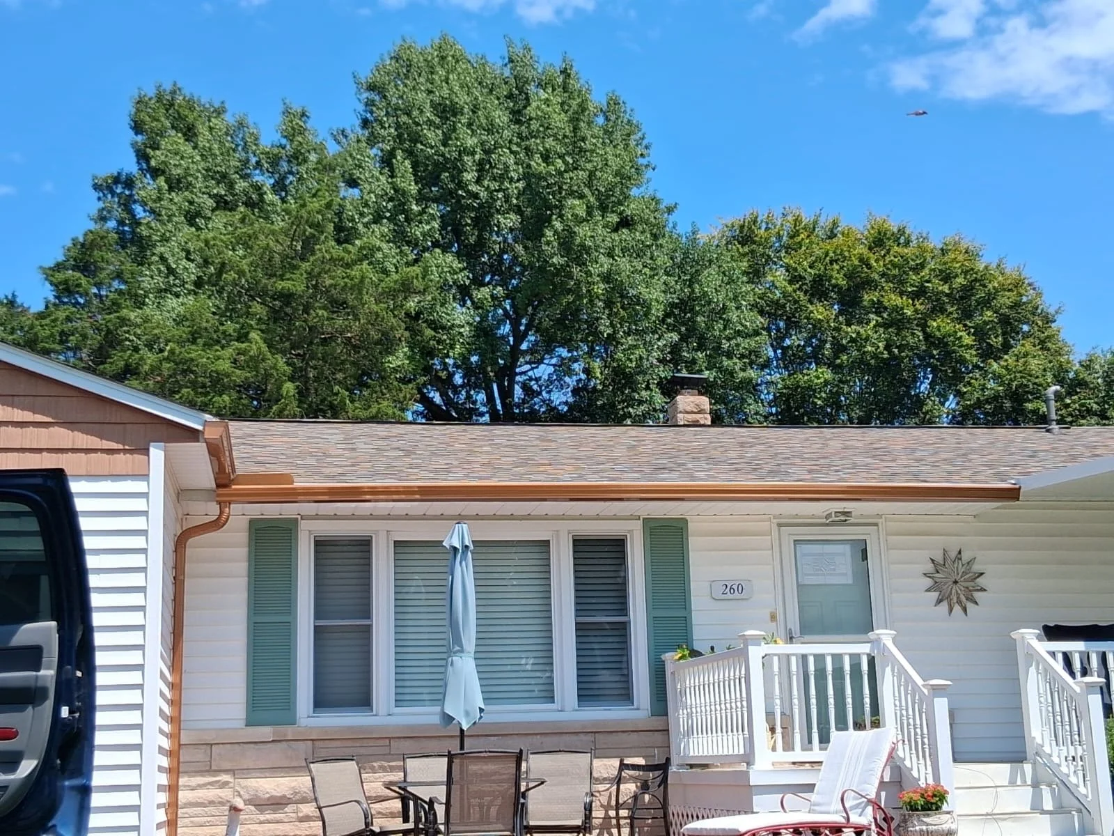 Front view of a house with white siding, a porch with white railing, patio furniture, and a door with the house number 260. There is a large star decoration on the wall, green shutters, and a brown shingled roof. Trees and a blue sky with some clouds