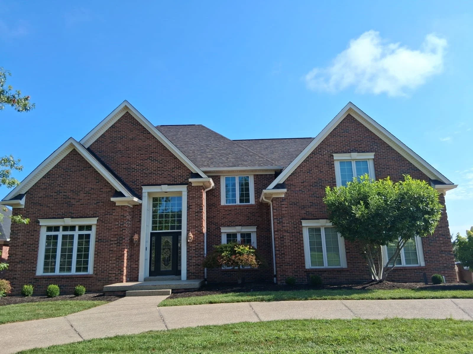 A large red brick house with white trim, multiple gabled roofs, and several windows, surrounded by a green lawn and bushes, against a blue sky with a few clouds.