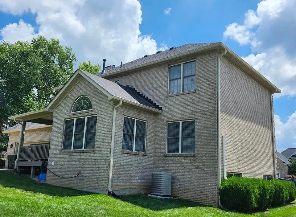 Exterior view of a beige brick two-story house with a high-pitched roof, multiple windows, a small wooden deck, and surrounding green lawn and bushes, under a partly cloudy sky.