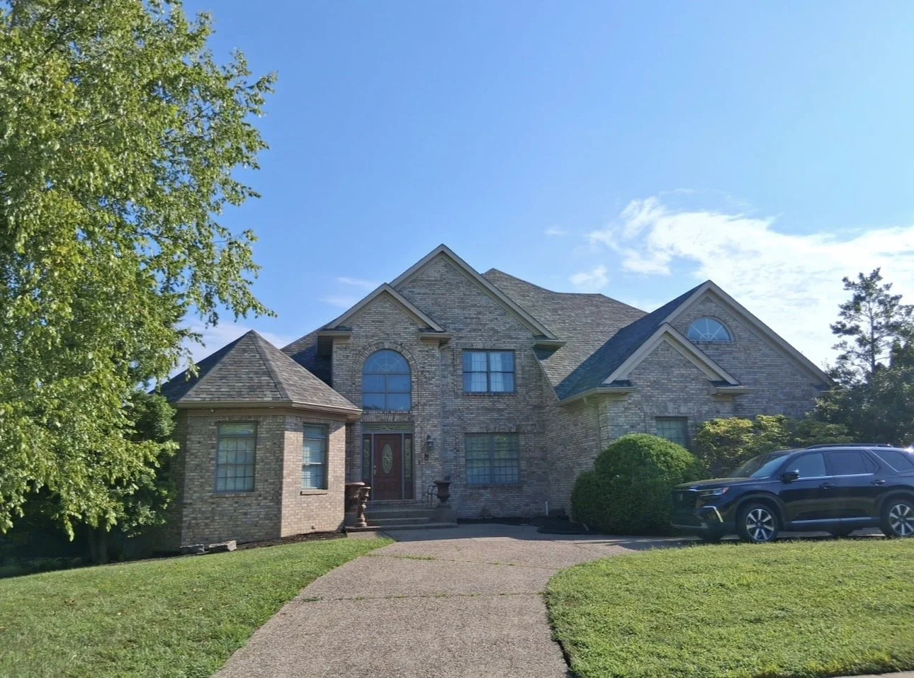 A large two-story brick house with a driveway and a black SUV parked on the right side. The house has multiple peaked roofs and arched windows, surrounded by green trees and bushes under a clear blue sky.