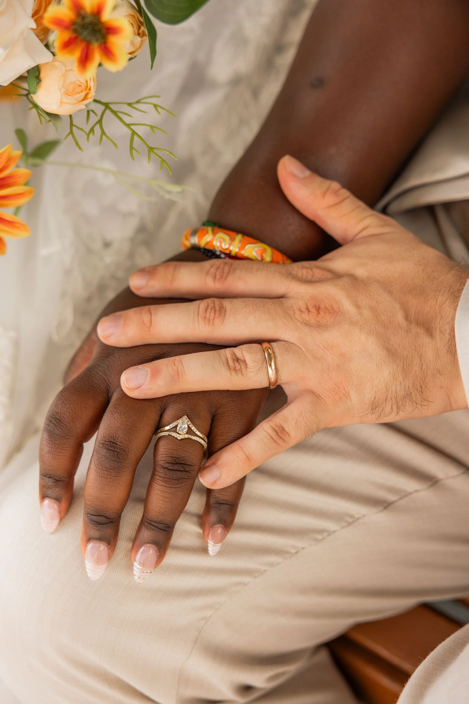 Close-up of two hands, one with dark skin and the other with light skin, gently holding each other. The dark-skinned hand displays an engagement ring and a wedding band, while the light-skinned hand wears a wedding band. A colorful bracelet adorns the dark-skinned wrist. Flowers are visible in the background.