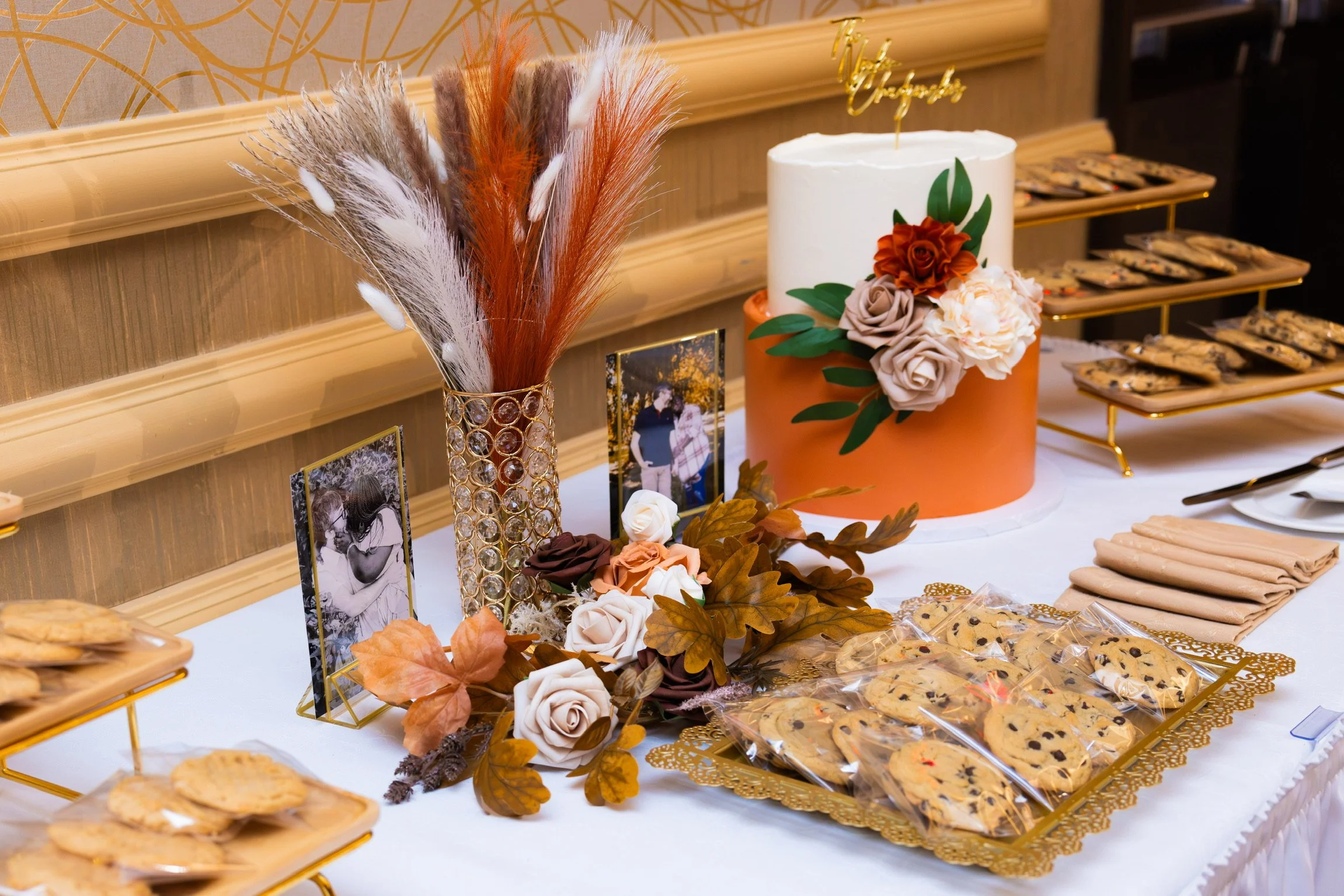 A decorated table with a large white cake adorned with flowers, framed photographs, a tall vase with pampas grass, and trays of cookies at a celebration or event.