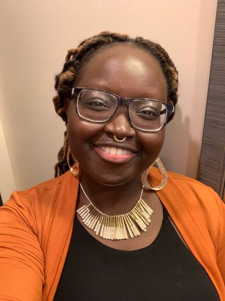 A woman with glasses, dreadlocked hair, and dark skin smiling. She is wearing large hoop earrings, a statement necklace, and an orange jacket over a black top. The background includes a pink wall and a wooden panel.