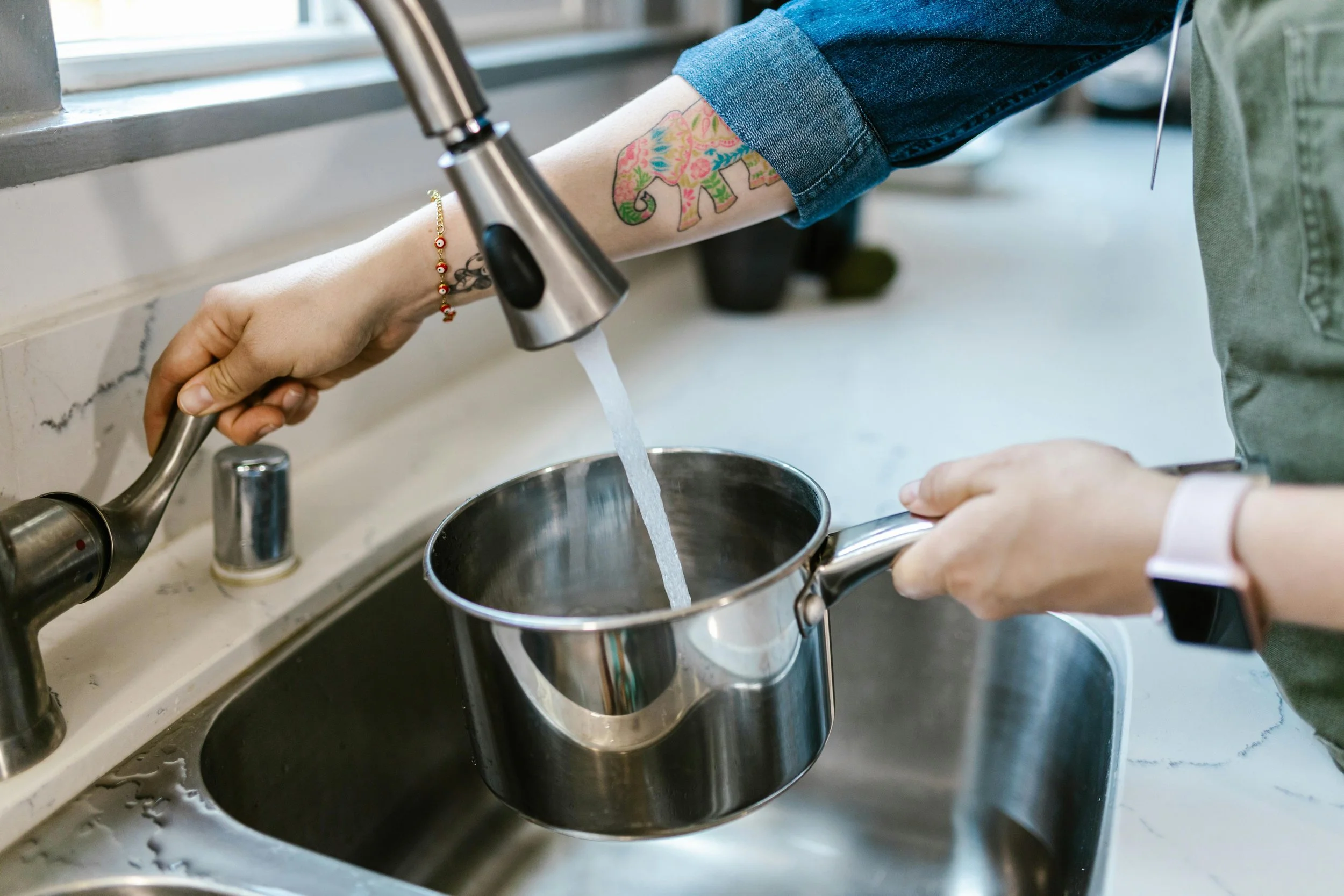 Person rinsing a pot under running water in a kitchen sink.