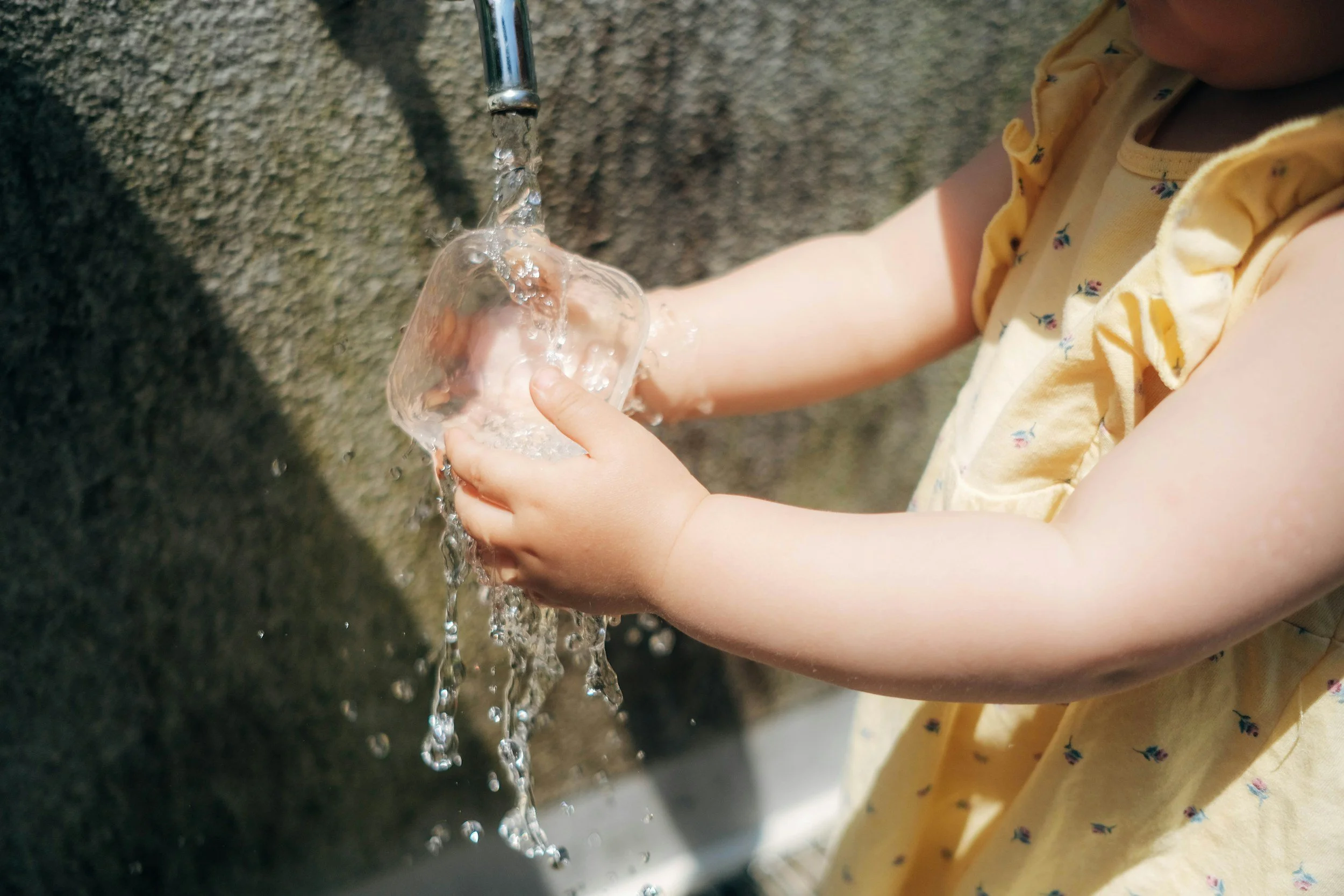 Child wearing a yellow dress washing a glass under running water.