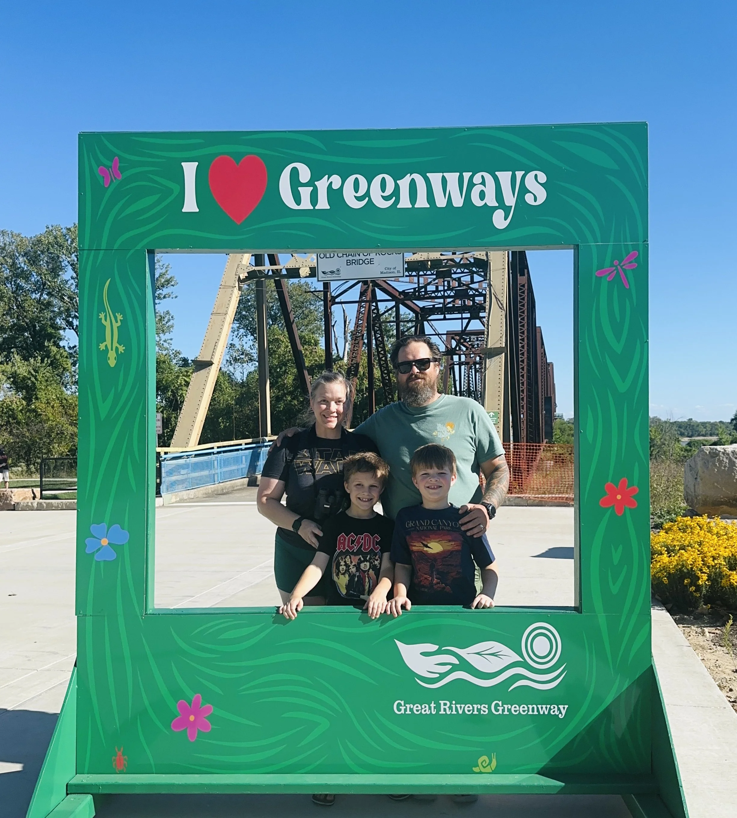 A family of four posing inside a green photo frame at the Great Rivers Greenway. The frame has the text 'I love Greenways' at the top and the logo of the greenway at the bottom. The family is standing on a paved pathway with a bridge in the background on a sunny day.