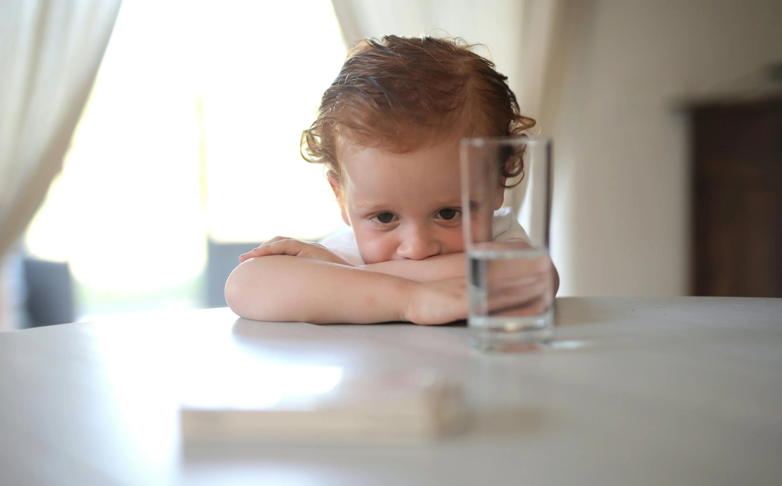 A young boy with curly red hair resting his head on his arms at a table, looking at a tall glass of water in front of him.