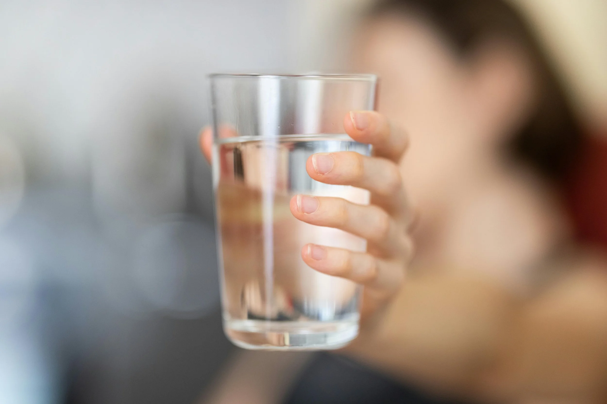 A person holding a clear glass of water with one hand, fingers wrapped around the glass, in focus. The background and person's face are blurred.