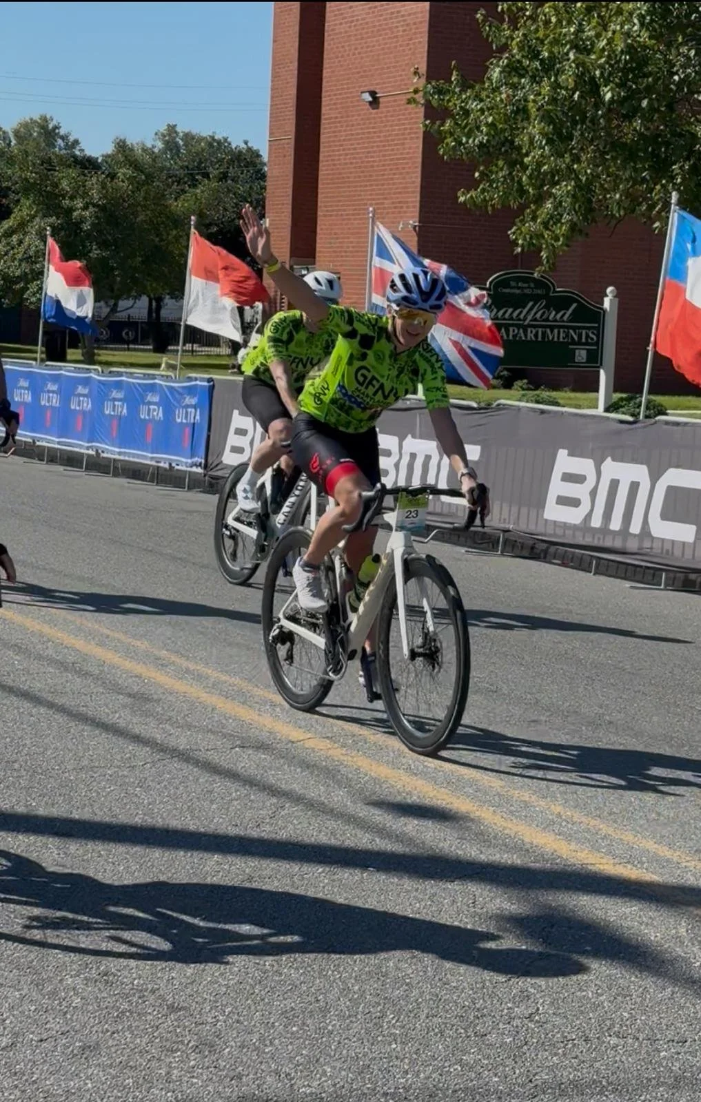 Yuliya Flynn is cycling in a GFNY race wearing a neon green and black jersey and a helmet, waving, on a road with banners and flags during a race event.