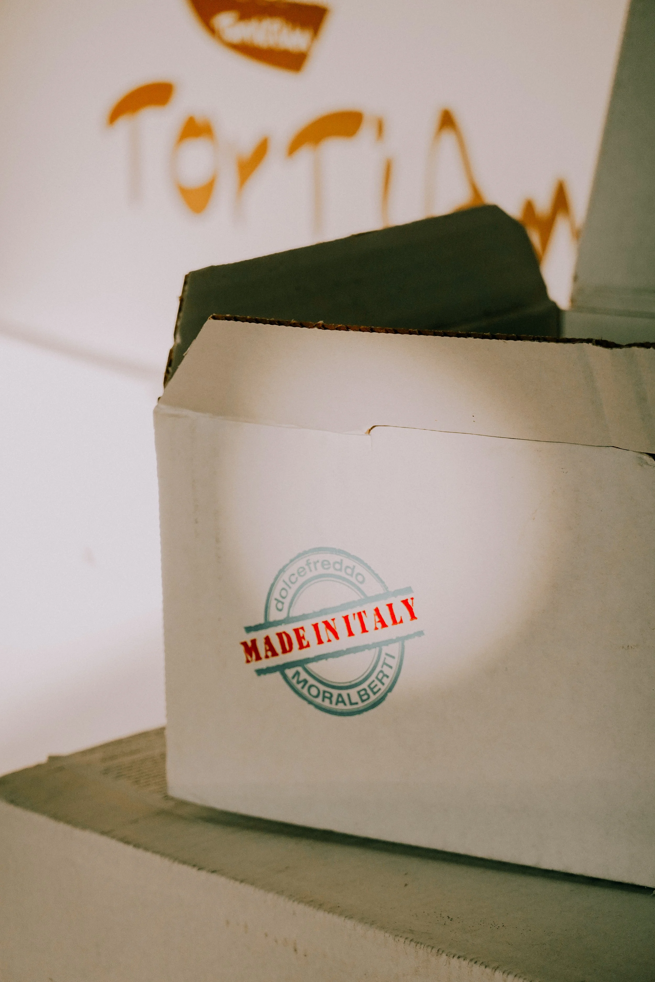 Close-up of a cardboard box labeled 'Made in Italy' with the logo of 'Dulce Freddo' and 'Moralbert' printed on it, placed on a surface with another box in the background.