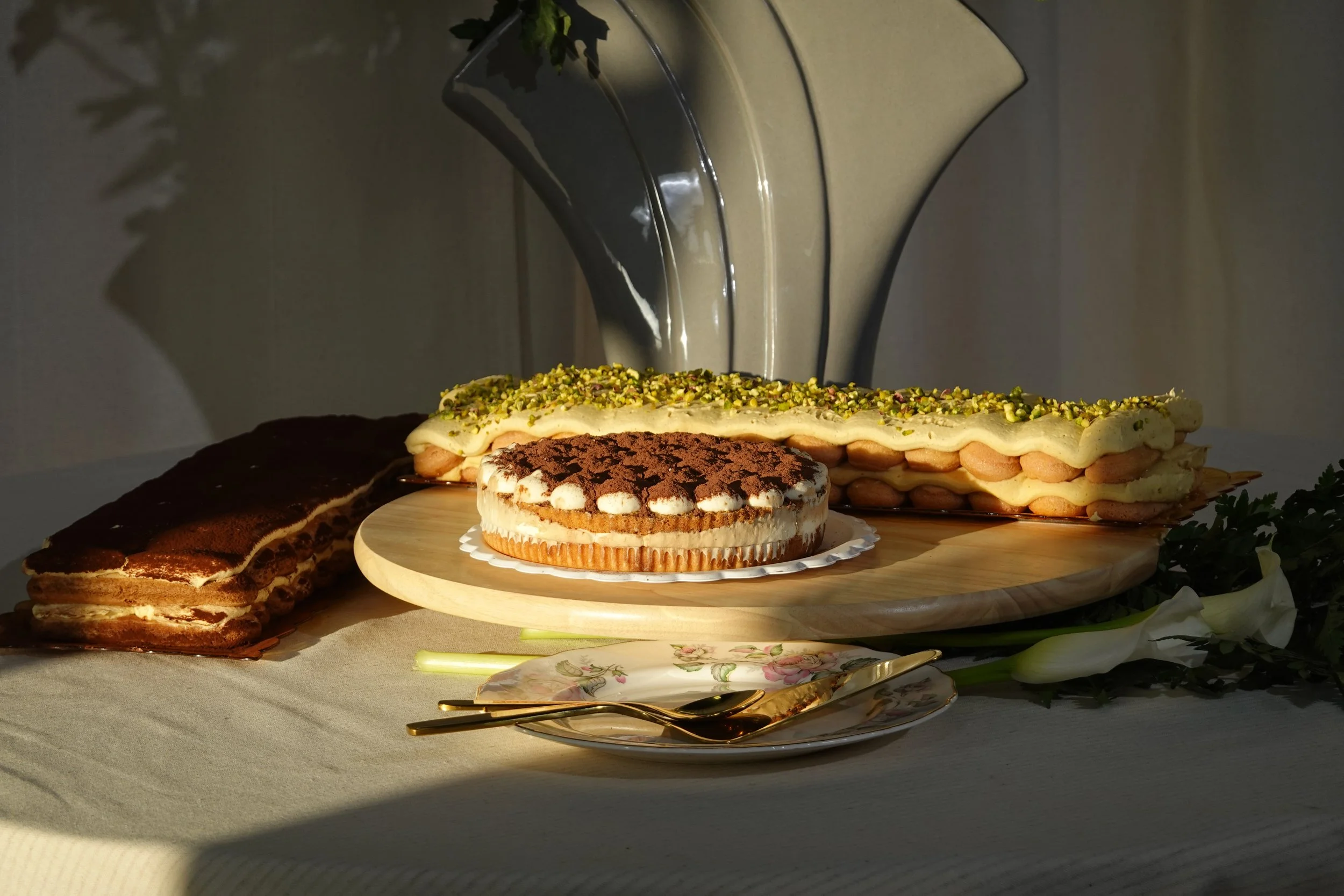 A selection of desserts including a large creme caramel, a chocolate and vanilla layered cake, and a pistachio pastry, displayed on a wooden platter with a gold fork and spoon, illuminated by a lamp, on a white tablecloth.