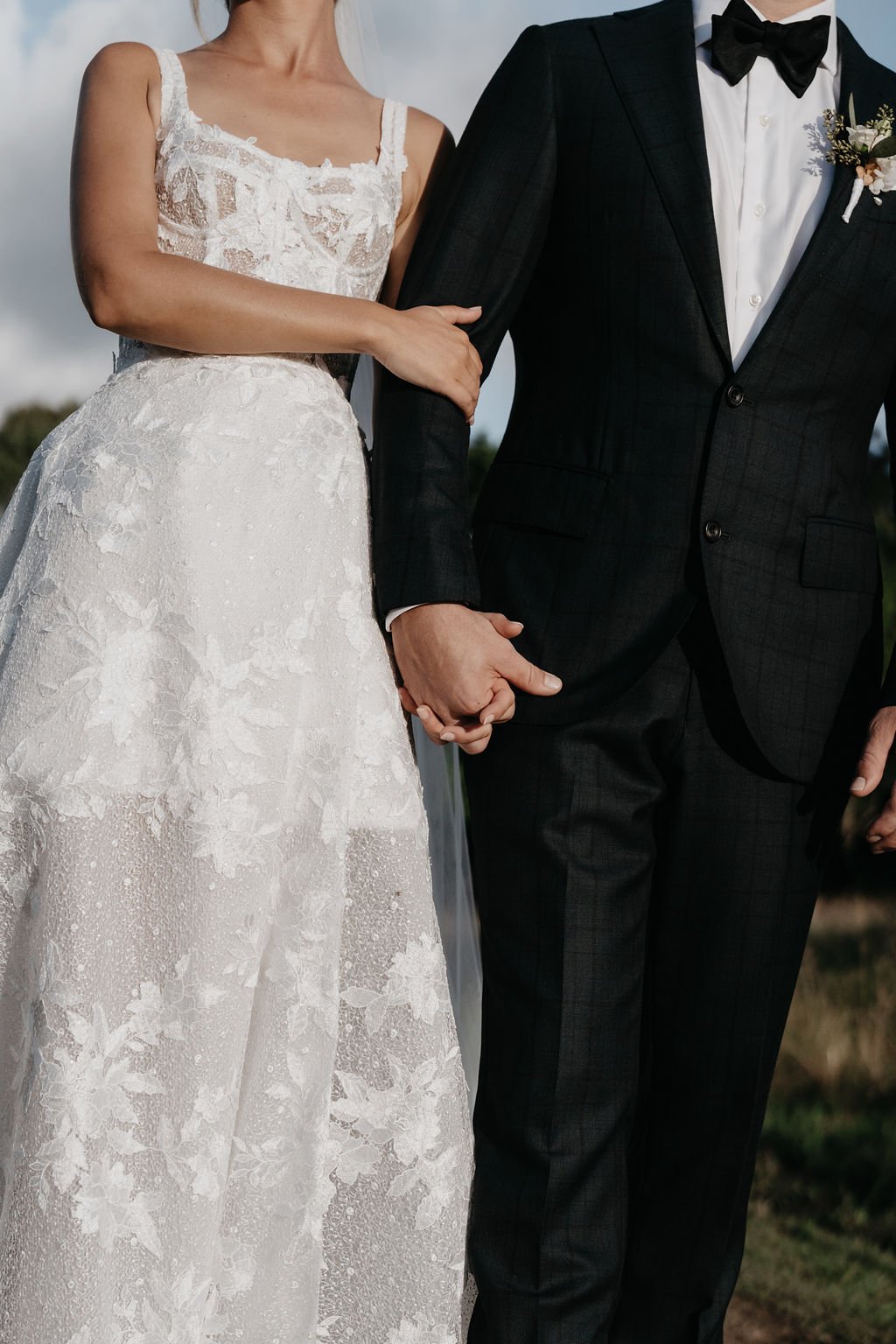 A bride and groom holding hands, dressed for wedding, outdoors with cloudy sky.