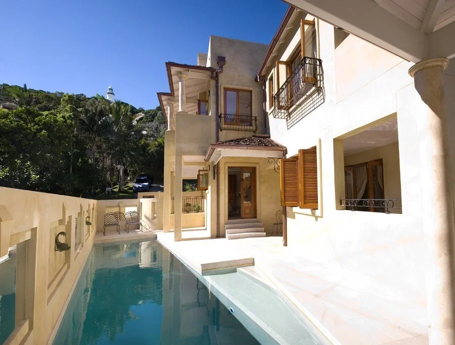 A house with a swimming pool in the foreground, beige exterior walls, wooden balcony shutters, and a hillside with trees and a lighthouse in the background.
