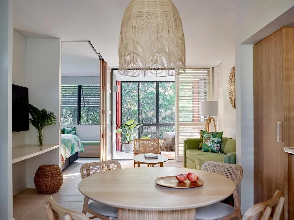 Open-concept living room and bedroom with natural light, wooden furniture, green cushions, tropical plants, and coastal decor.