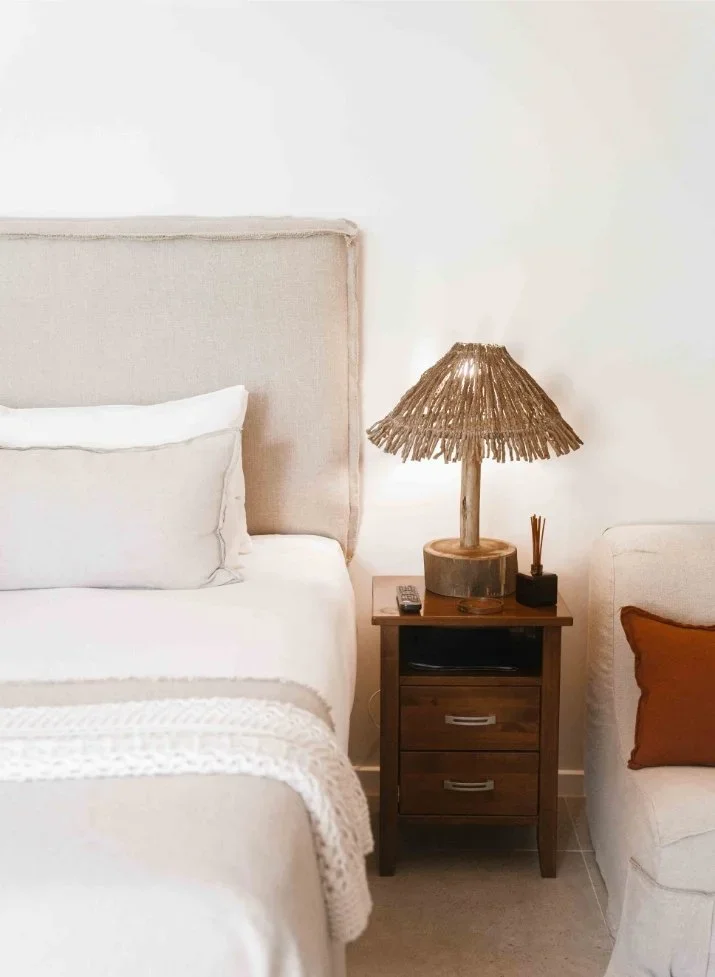 A bedroom corner with a beige upholstered bed, white pillows, a wooden nightstand with a rustic lampshade made of natural fibers, a remote control, and a small reed diffuser. There is also a white sofa with an orange pillow next to the nightstand.