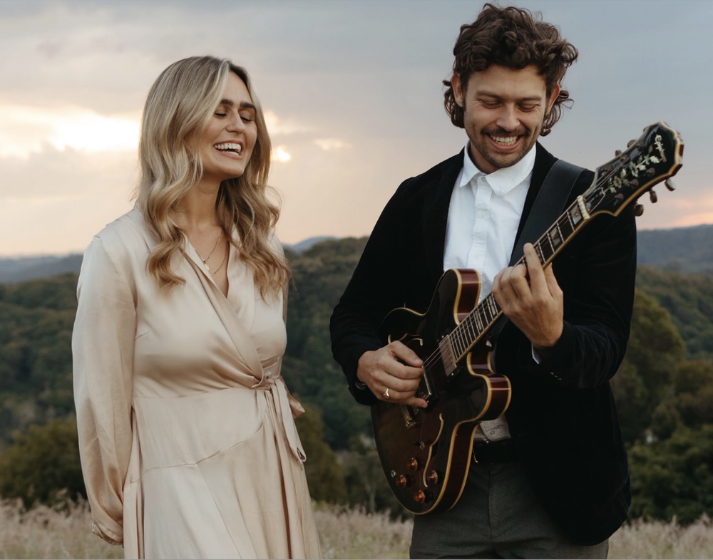 A man playing an electric guitar outdoors with a woman standing nearby, both smiling and enjoying the moment during sunset or late afternoon.