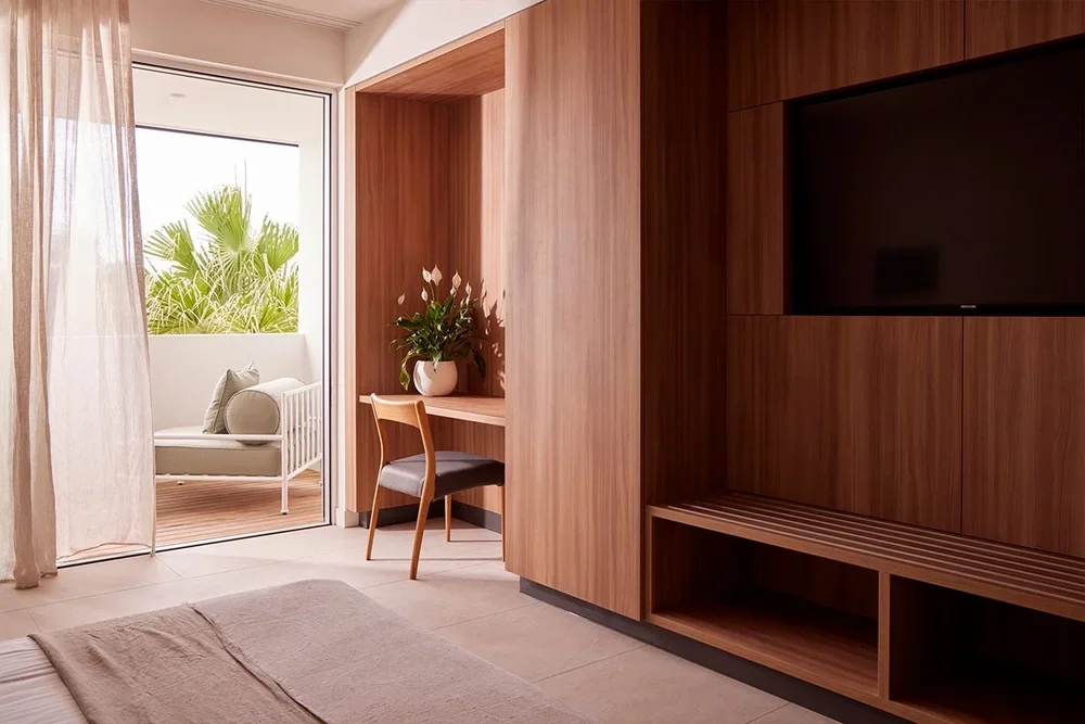 A cozy hotel room with a bed, wooden entertainment center with TV, small workspace with a plant, and a patio with seating and greenery visible through the glass door.