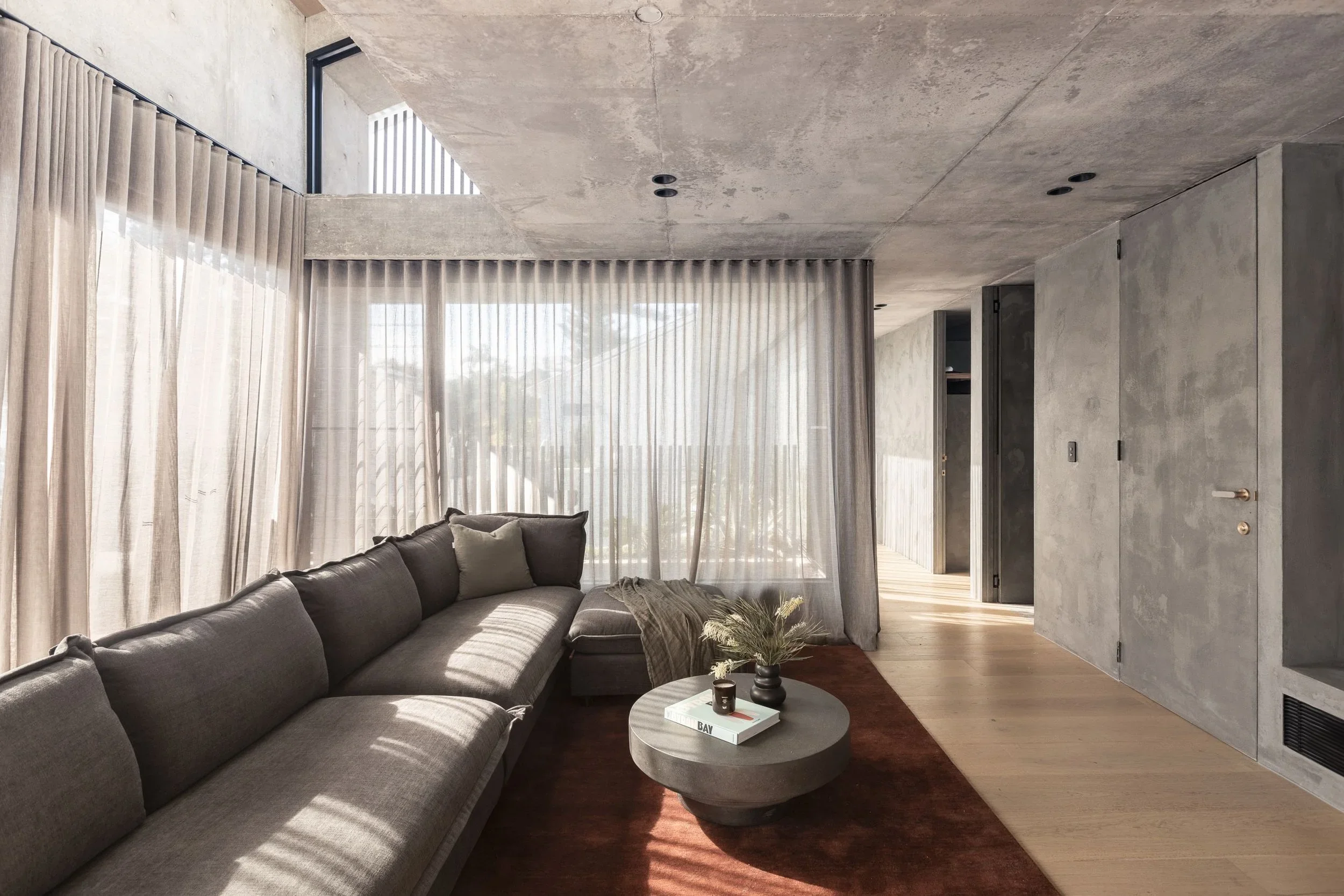 Modern living room with concrete ceilings and walls, large windows with sheer curtains, a dark gray sectional sofa with beige and gray pillows, a round coffee table with a plant, books, and decor, and hardwood flooring.