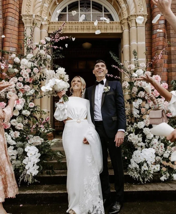 A bride and groom smiling happily at their wedding celebration, standing in front of a church entrance adorned with flowers. The bride is holding a bouquet of flowers, and guests are throwing flower petals.