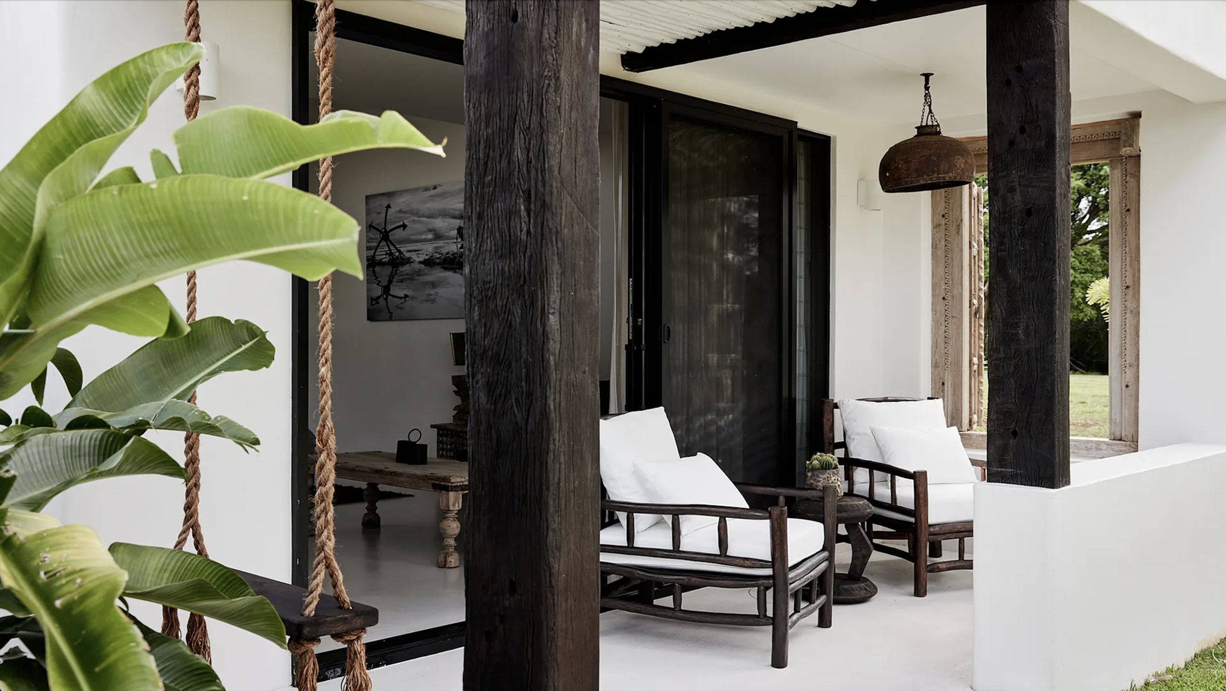 Porch area with wooden columns, white furniture, large green plants, and an open view of greenery outside.