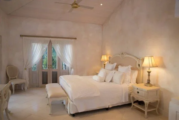 A bedroom with a large bed, nightstands, and lamps, with French doors with white curtains leading outside.