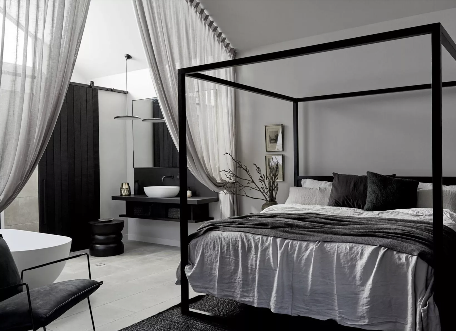 Modern bedroom with four-poster bed, white bedding, black and white pillows, and a side table with a vase. In the background, there is a black and white bathroom with curtains, a sink, and framed artwork on the wall.