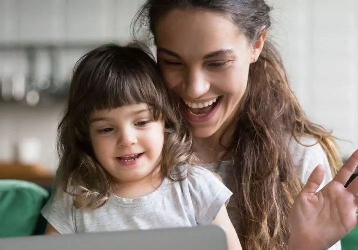 A young woman and a little girl smiling and looking at a laptop together in a room with a blurred background.