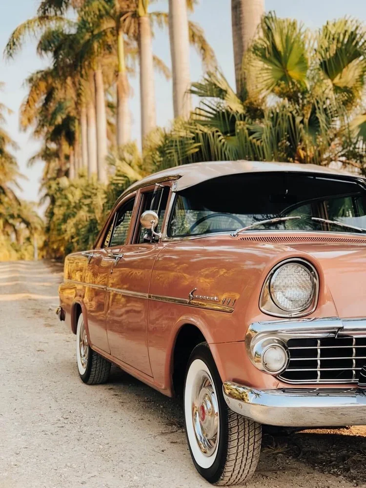 Vintage peach-colored car with a white roof parked on a sandy road, surrounded by tall palm trees and lush greenery.