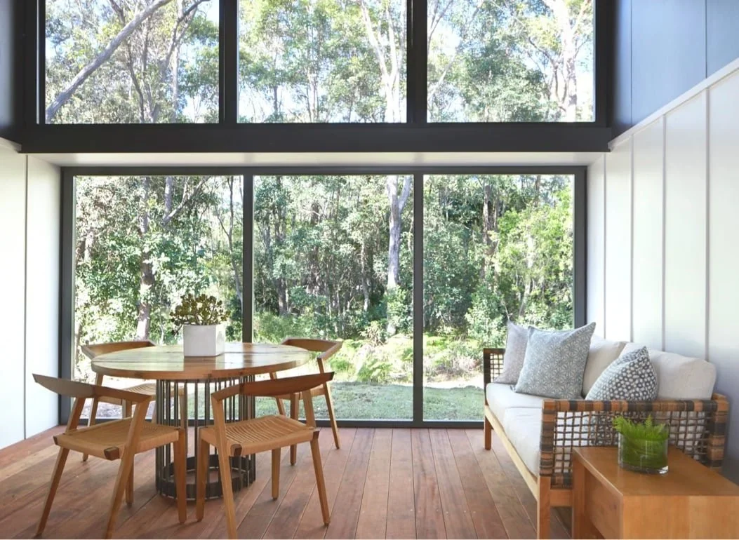 Bright room with large floor-to-ceiling windows showing trees outside. Wooden dining table with four chairs, a white sofa with patterned pillows, and a small wooden side table with a plant.