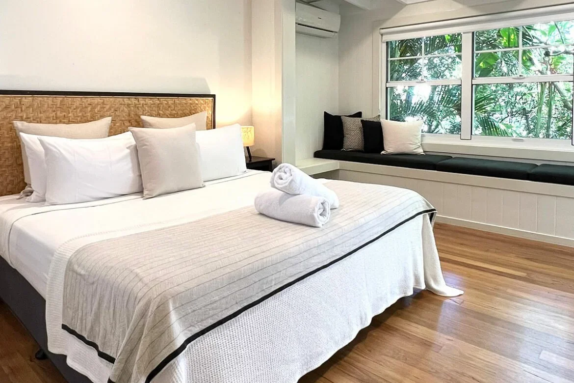Comfortable bedroom with a bed featuring white linens, three pillows, and two rolled towels on top, beside a window with lush greenery outside, and a cozy window seat with pillows.