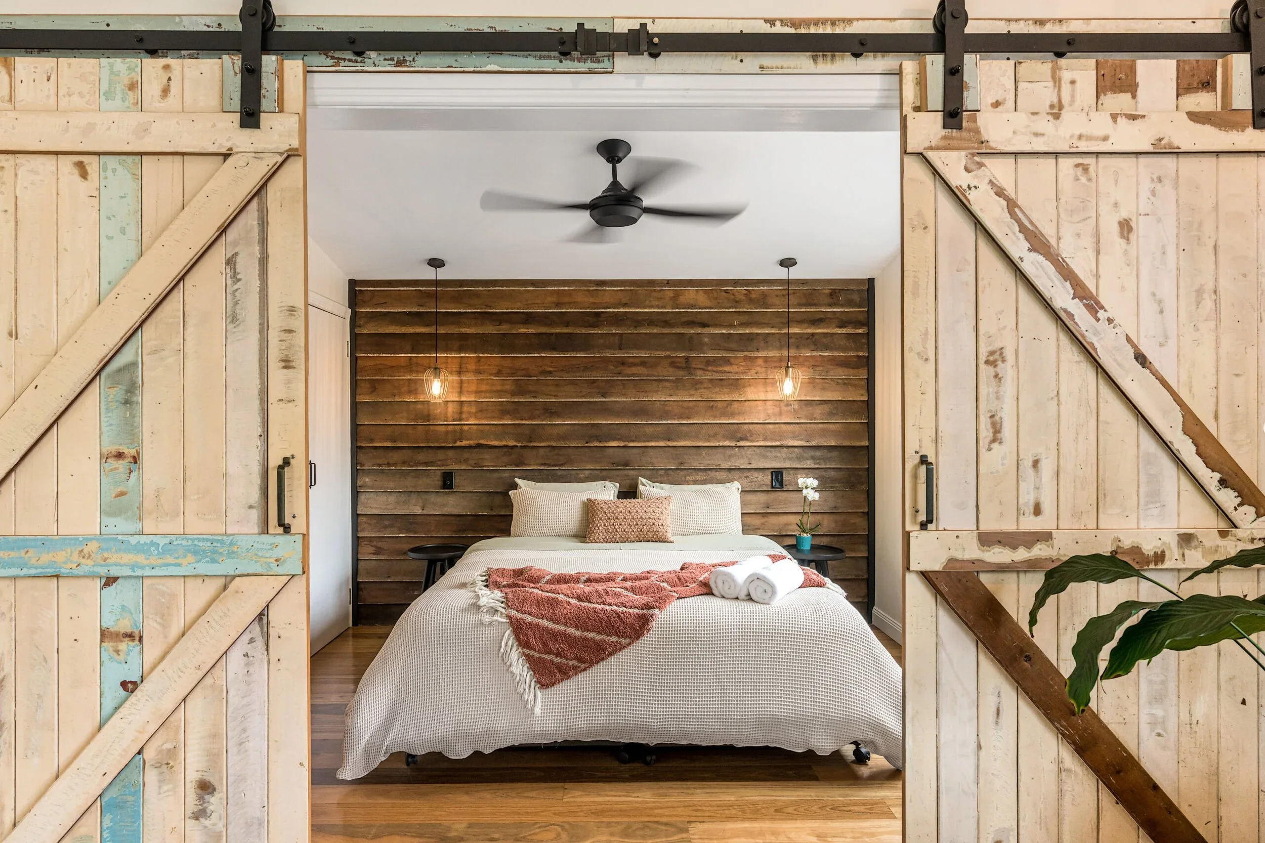 View of a bedroom through partially open rustic sliding barn doors made of distressed wood with some peeling paint. The bedroom has a bed with light-colored bedding, a throw blanket, pillows, a small side table, and pendant lights hanging from a wooden-paneled ceiling. A ceiling fan is also visible.