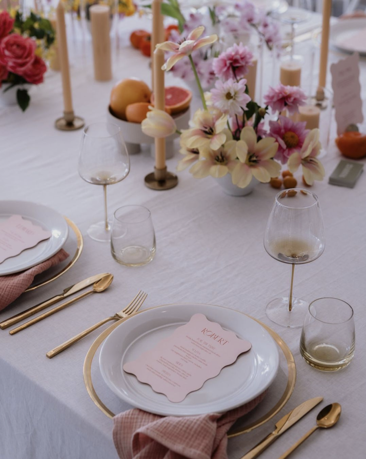 Elegant table setting for a celebration with white plates, pink napkins, gold utensils, and pink-themed menus. Decor includes pink and white flowers in a vase, pink candles, and fruit such as grapefruit, oranges, and apricots.