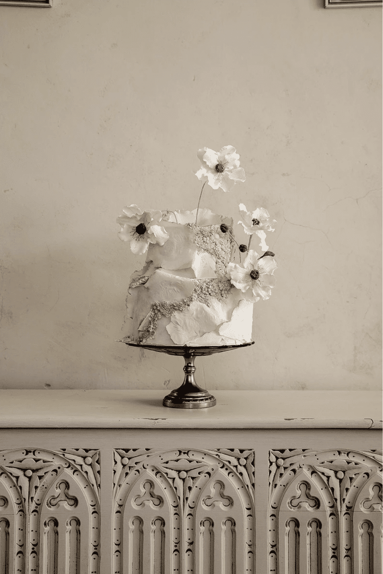 A decorative cake with white flowers on top, placed on a silver cake stand, on a white ornate surface against a plain wall.