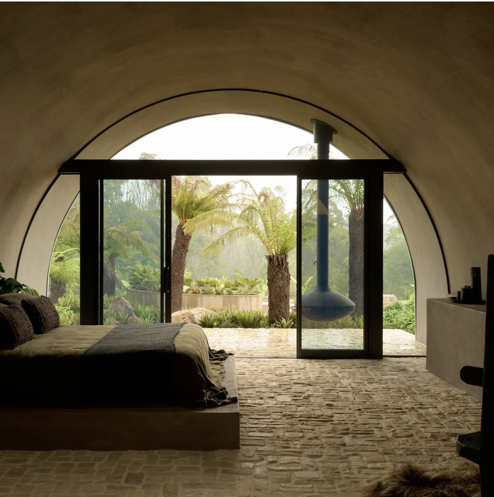 A cozy bedroom with a large arched window showing lush greenery and palm trees outside. The room has a bed with dark pillows and blanket, brick flooring, and minimal decor.