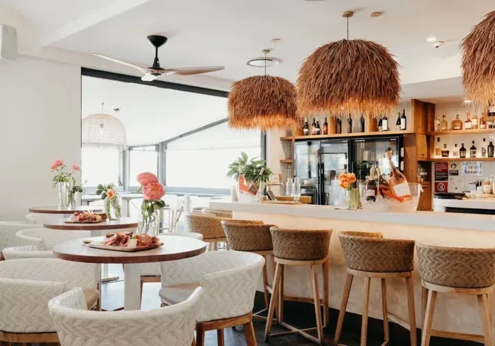 A bright, modern restaurant interior with round tables set with plates, glasses, and pink flowers. There is a bar with bar stools, bottles, and decorative hanging light fixtures.