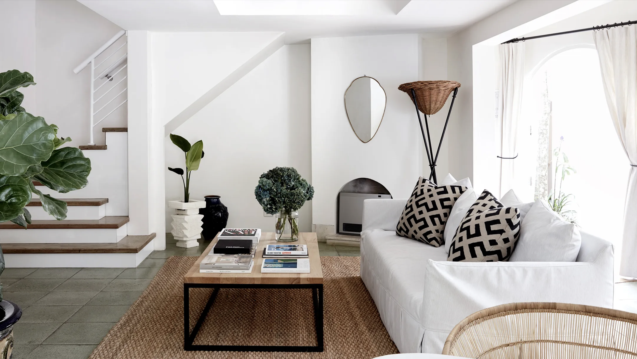Living room with white walls, a white sofa with black geometric pillows, a wooden coffee table with magazines, green plants, and a wicker basket lamp near window with white curtains.