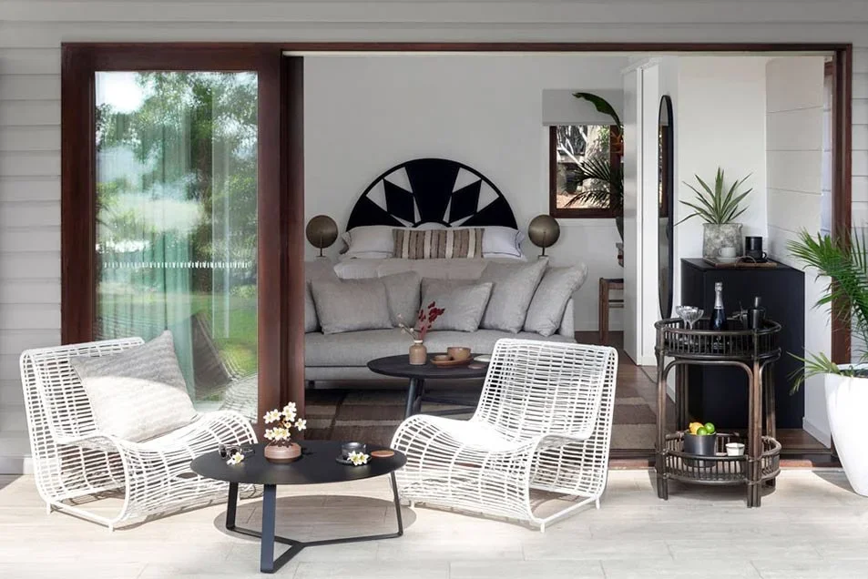 Modern open-concept living space with white wicker chairs, black coffee table, and a cozy bedroom with a bed and decorative pillows visible through an open wall.
