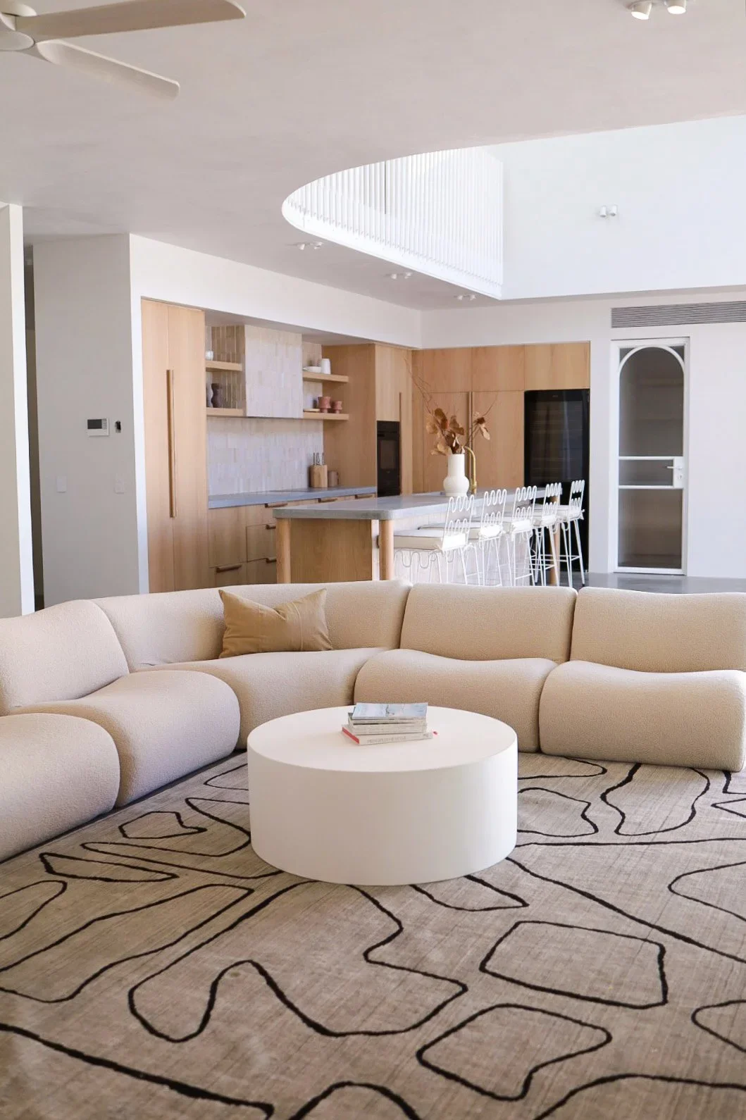 Modern living room with a beige sectional sofa, a round white coffee table, a patterned area rug, and an open kitchen with a breakfast bar and white chairs.
