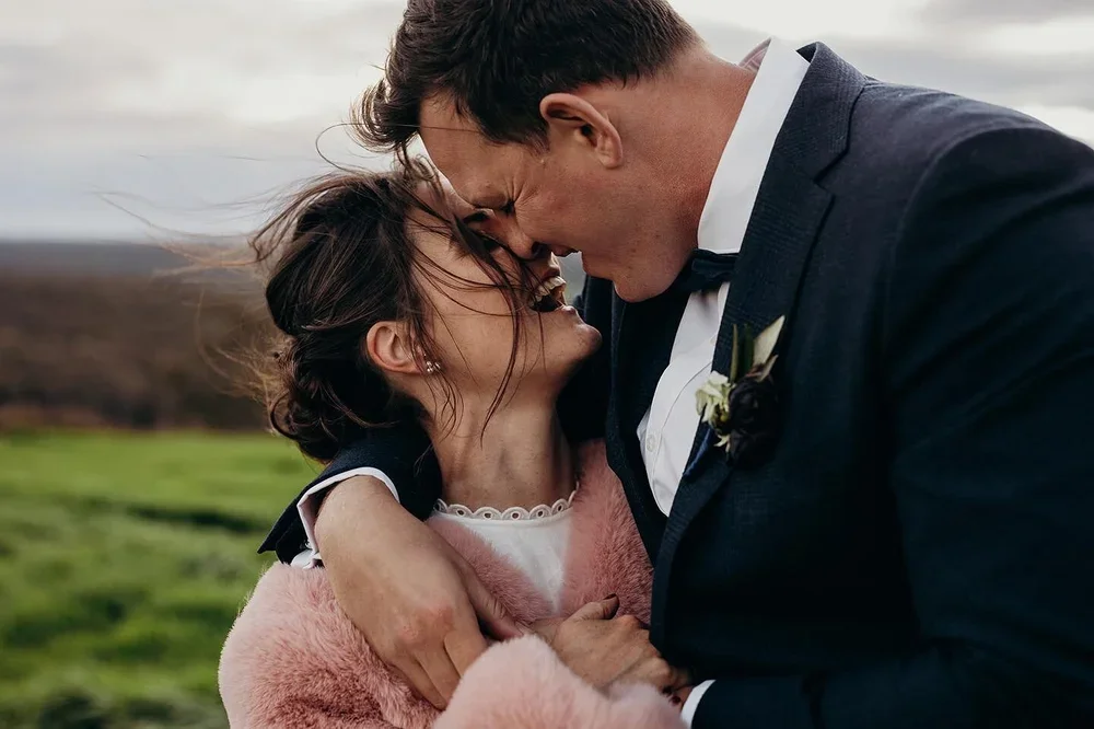 A couple dressed in wedding attire embracing outdoors, with their foreheads touching and smiling. The woman is wearing a pink coat, and the man is in a dark suit with a boutonniere, against a natural landscape background.