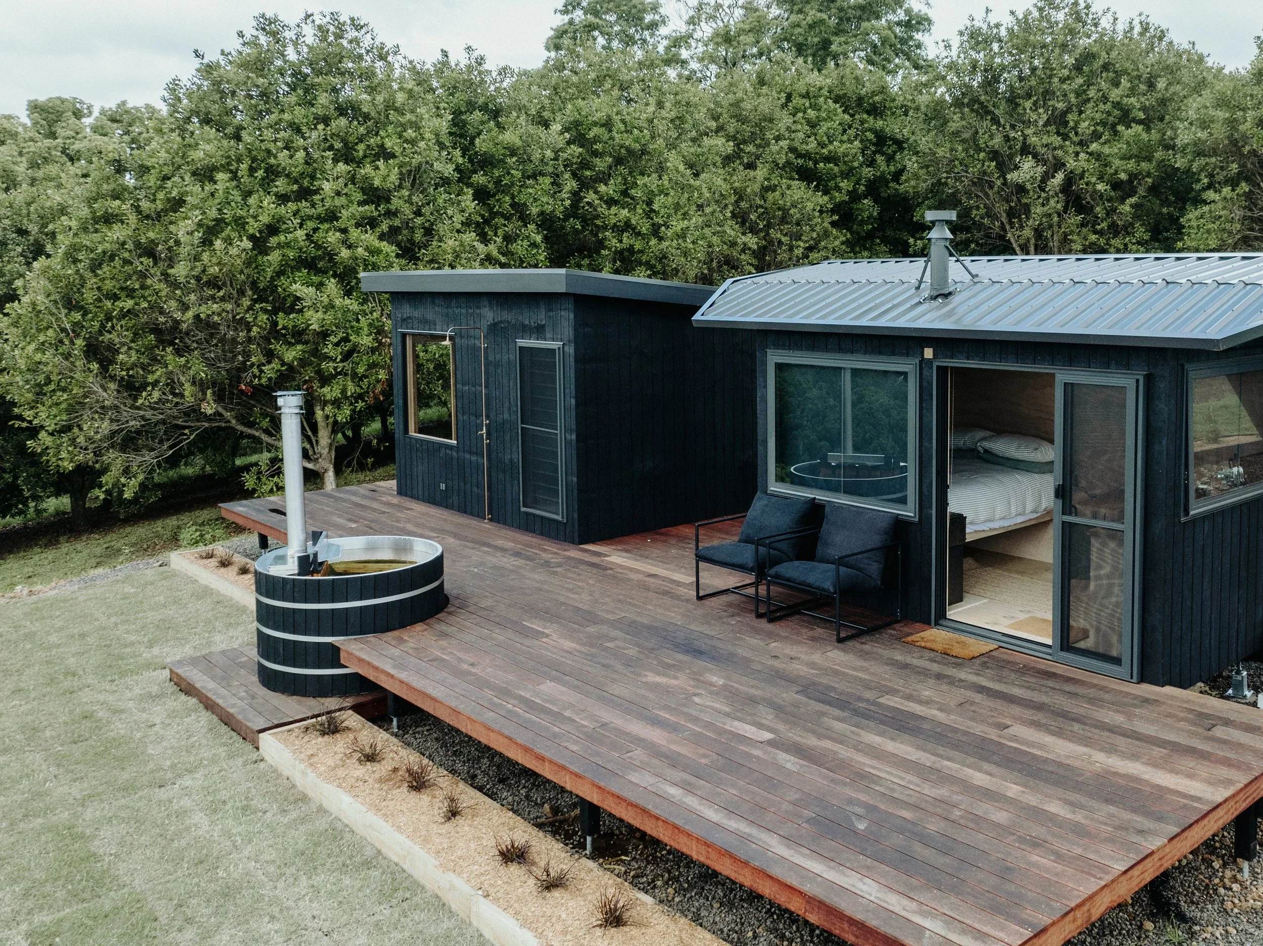 Modern tiny house with black exterior, large windows, and a metal roof, situated on a wooden deck with outdoor seating. Surrounding lush green trees and a cloudy sky.