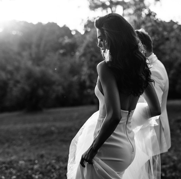 Black and white photo of a woman in a wedding dress holding the skirt with her hand, standing outdoors with trees in the background.