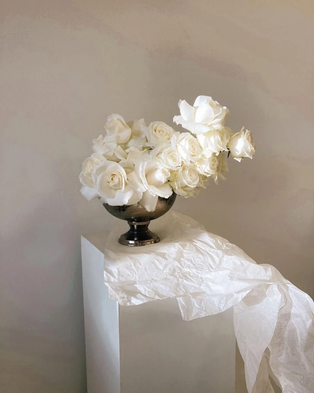 White roses in a dark metal vase placed on a white pedestal with crumpled white paper draped over it, against a plain beige wall.
